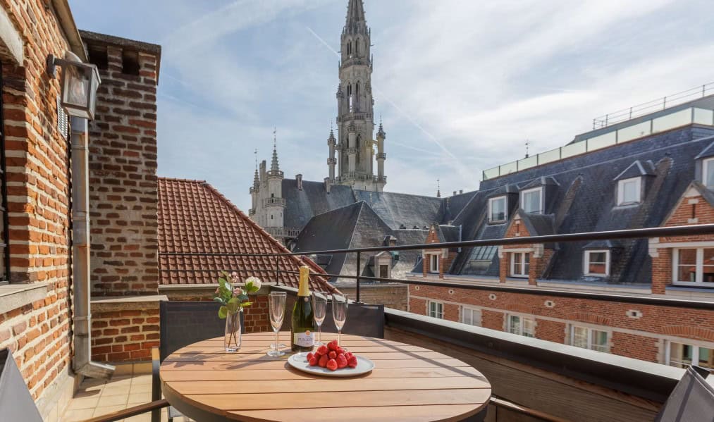 A round wooden table with champagne, glasses, and strawberries sits on the Grand Place Terrace overlooking brick rooftops and a tall church spire.