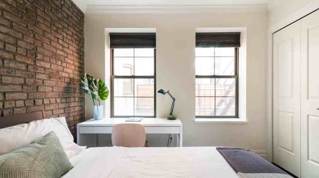Bright modern bedroom with brick wall, desk, and large windows.