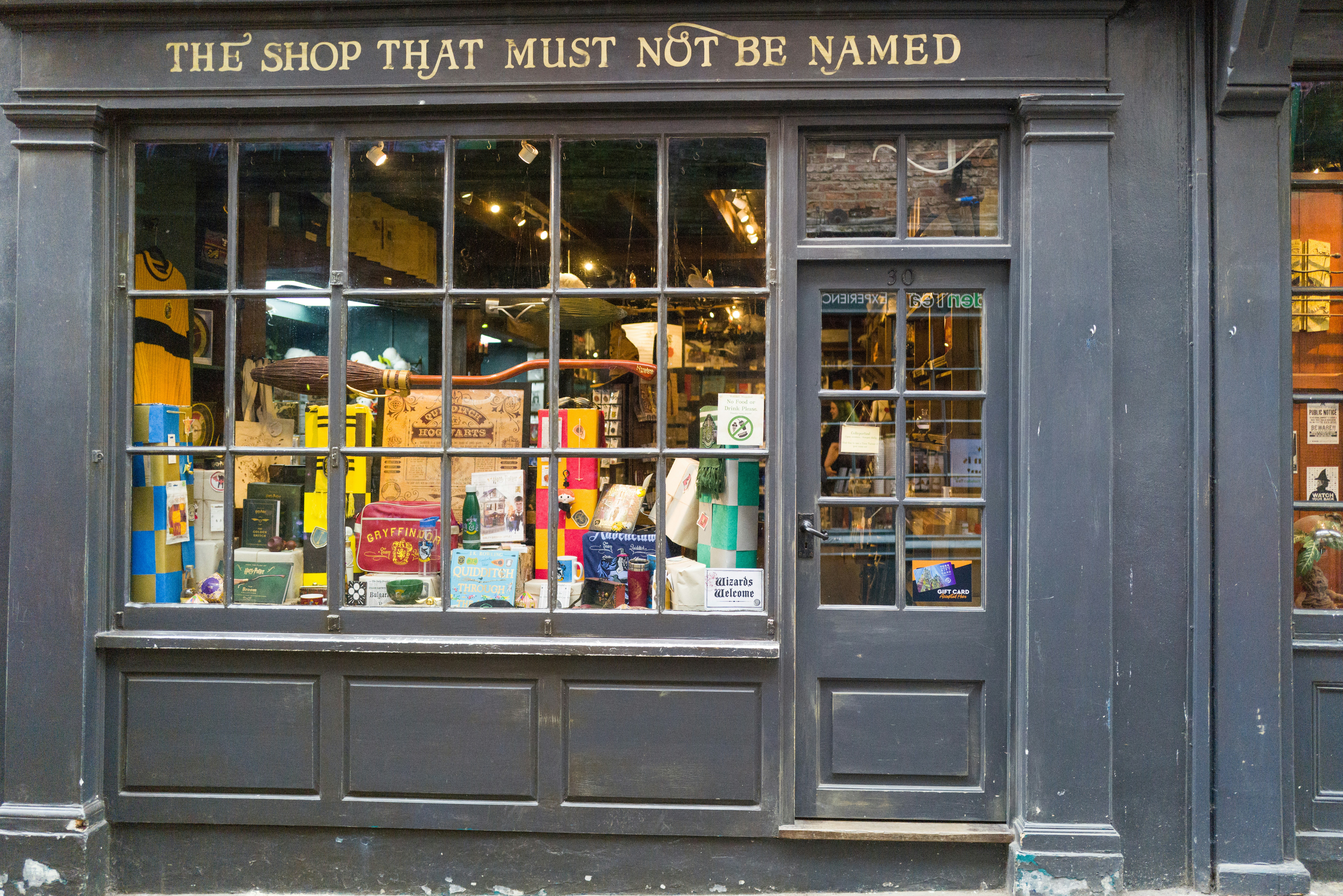 A storefront with the sign “The Shop That Must Not Be Named,” featuring large display windows and a glass door with colorful merchandise inside.