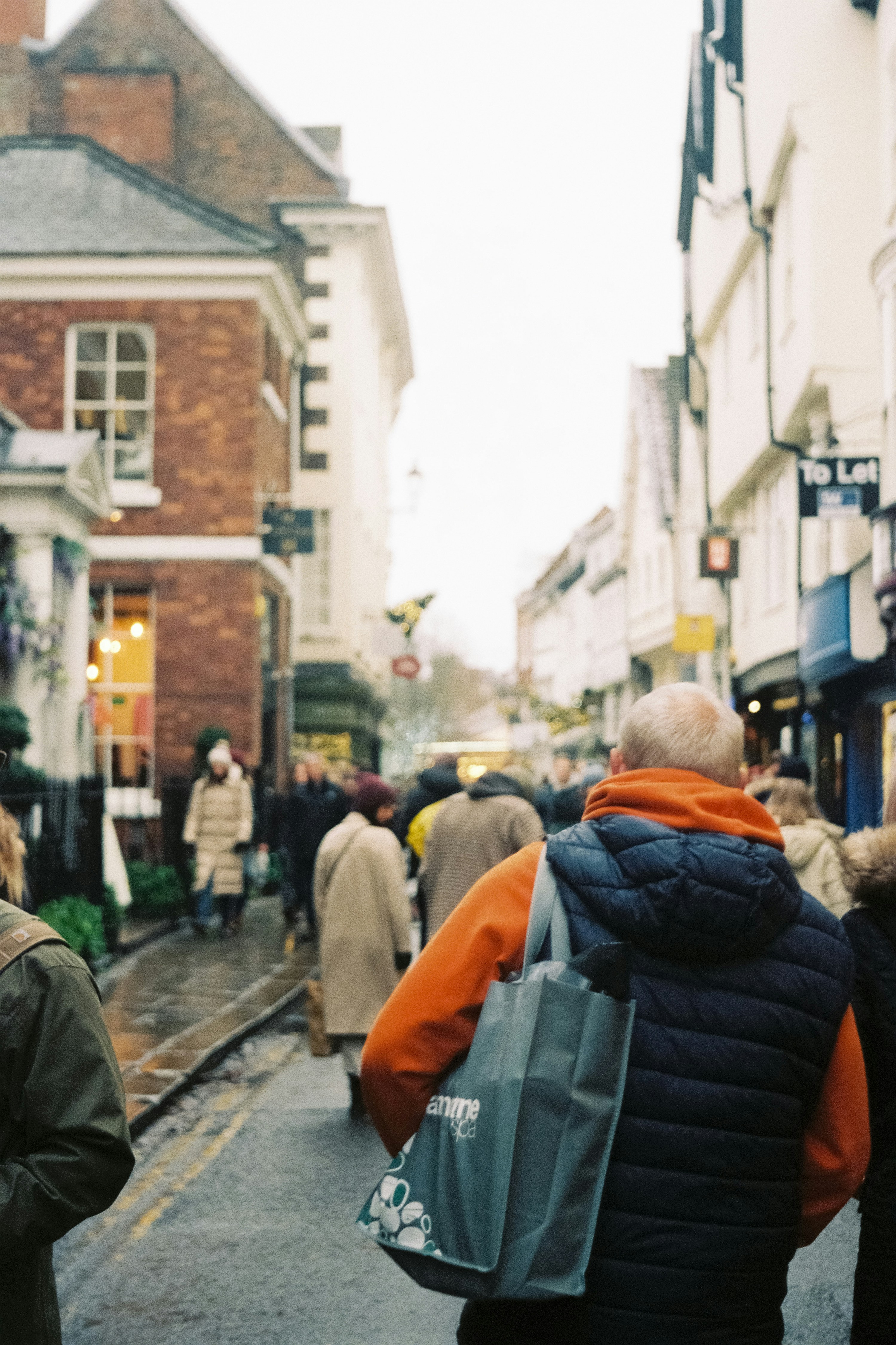 A man in an orange hoodie and navy vest carries shopping bags along a crowded wet street lined with shops and pedestrians.