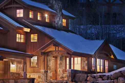 Cozy mountain cabin with warm lights and snow-covered roof at night.