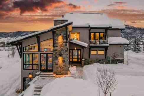 Modern mountain cabin with large windows in a snowy landscape at sunset.