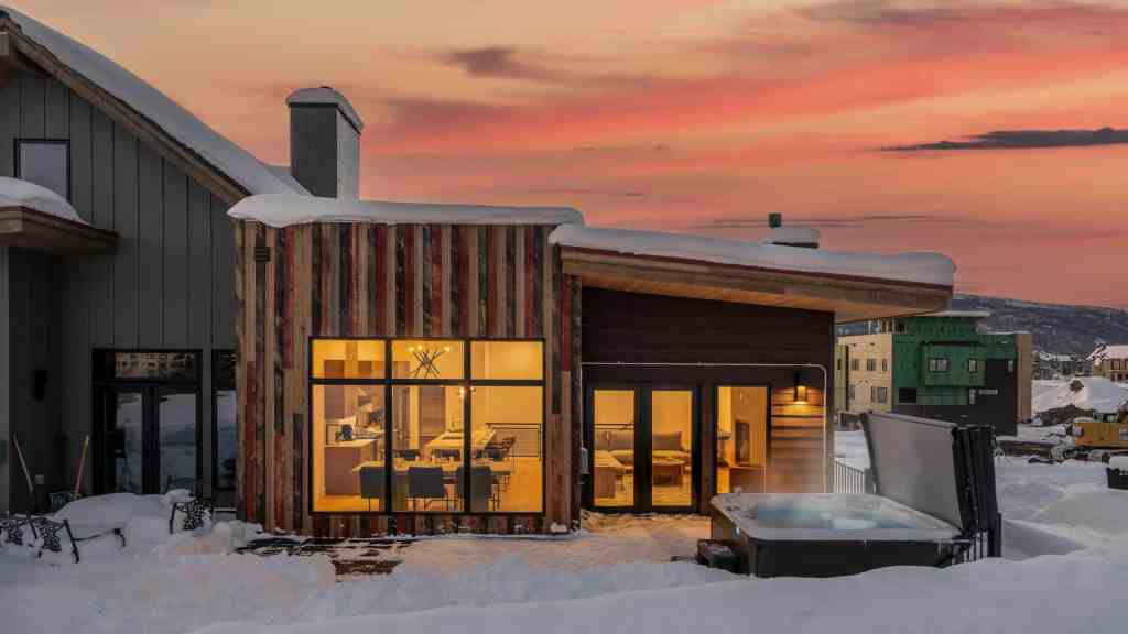 Modern alpine villa with large windows and hot tub in snowy landscape.