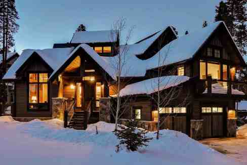 Luxury chalet with snow-covered roof and warm interior lights at dusk.