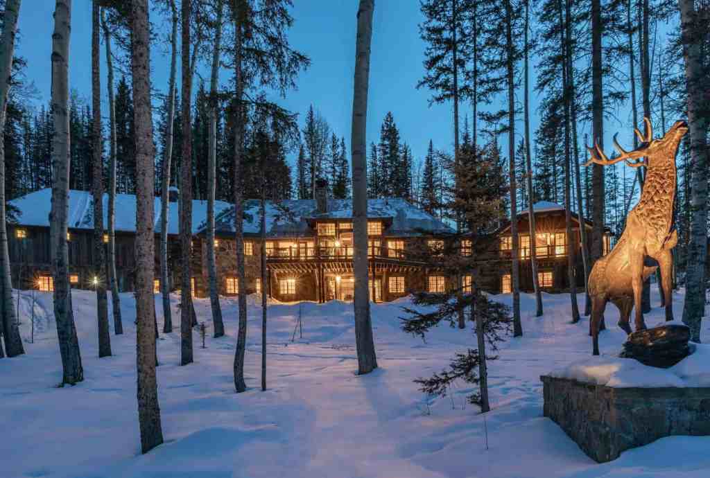 Snow-covered lodge with a large elk statue in a forest at dusk.
