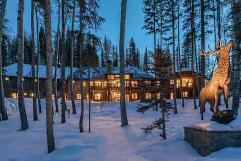 Snow-covered lodge with a large elk statue in a forest at dusk.