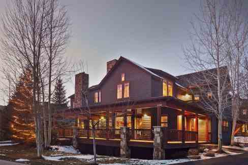Cozy wooden lodge with porch in winter, snow-covered landscape, leafless trees, warm lighting.