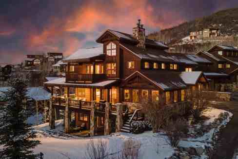 Charming mountain cabin with warm lights in a snowy setting at sunset.