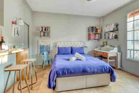 Bright bedroom with blue bedspread, bookshelves, and large windows.