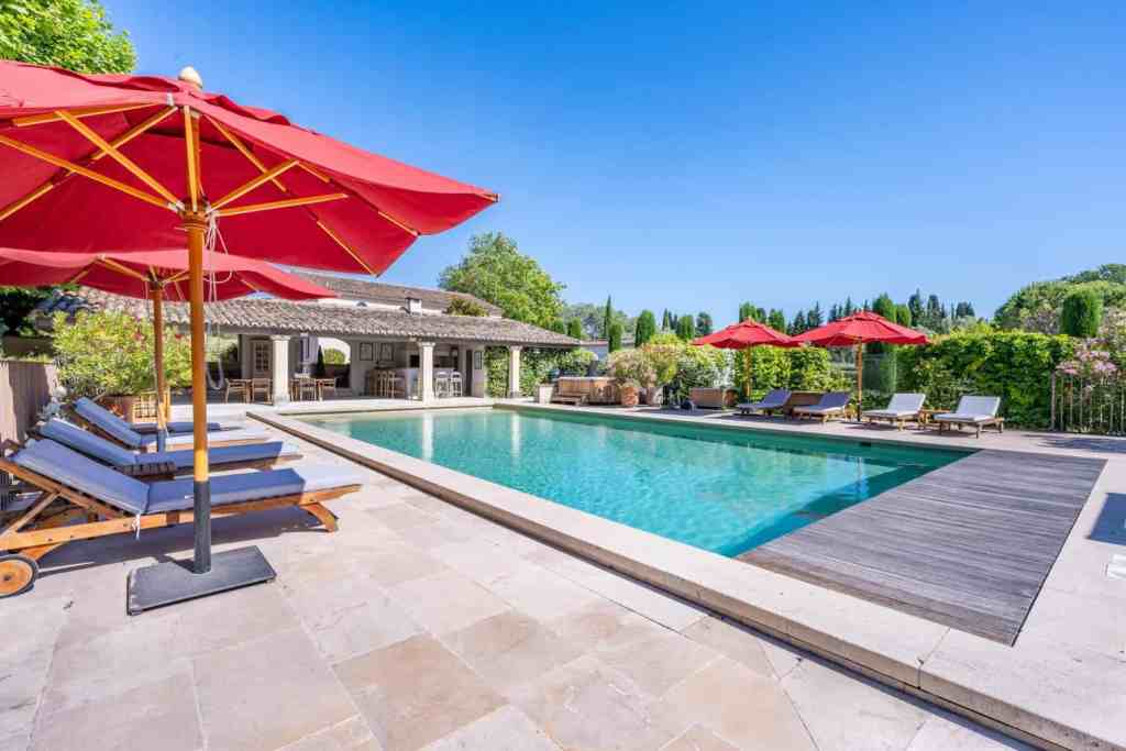 Elegant outdoor pool with sun loungers and red umbrellas in a sunny setting.
