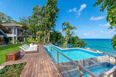 Elegant infinity pool overlooking the ocean with lush greenery and modern villa in the background.